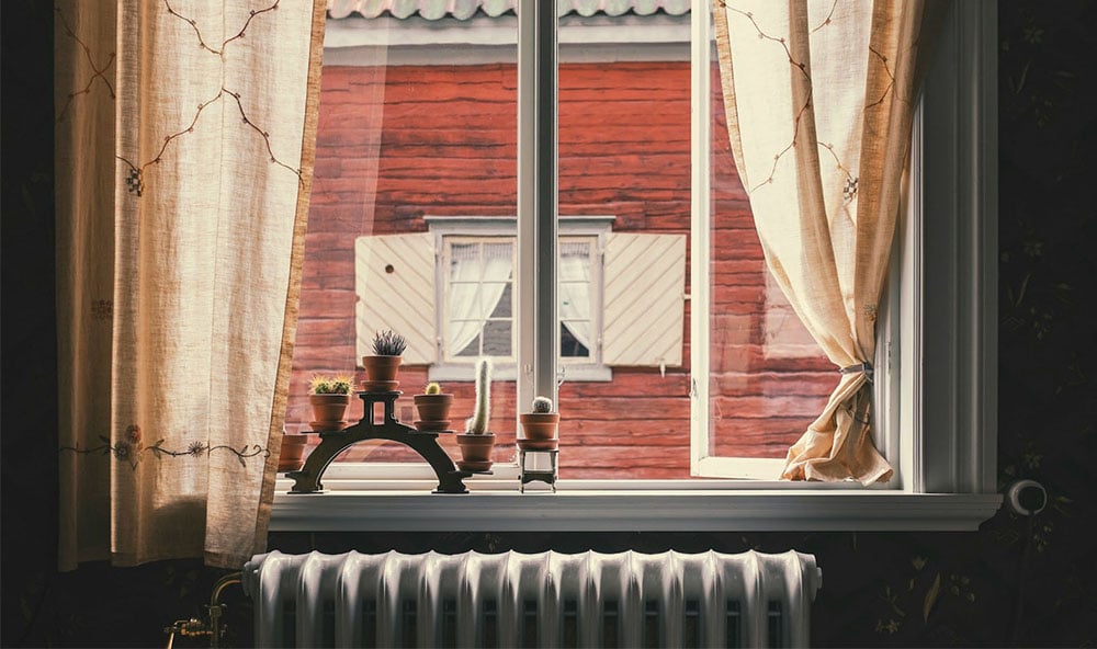 Öppet fönster med orangea gardiner med utsikt mot ett gammat rött hus.